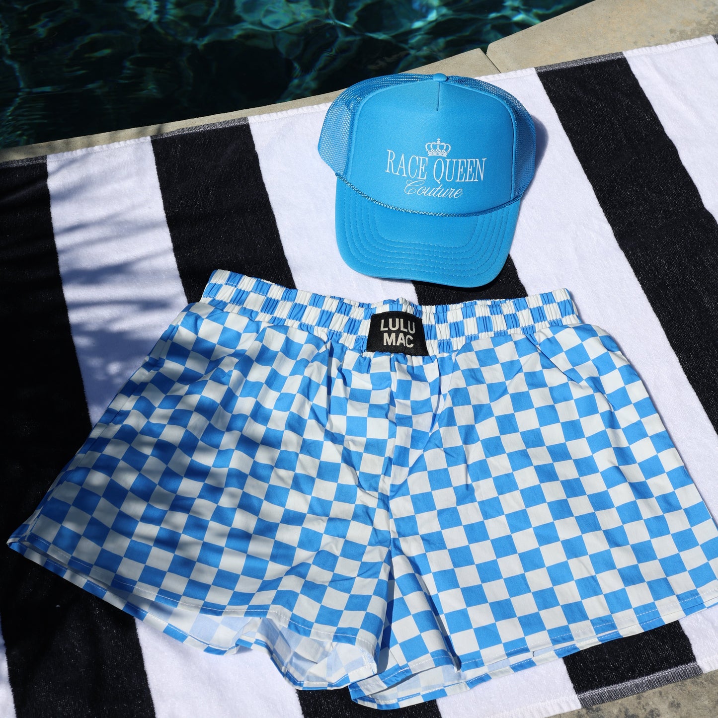 Blue checkered shorts and blue cap on a striped towel with water in the background