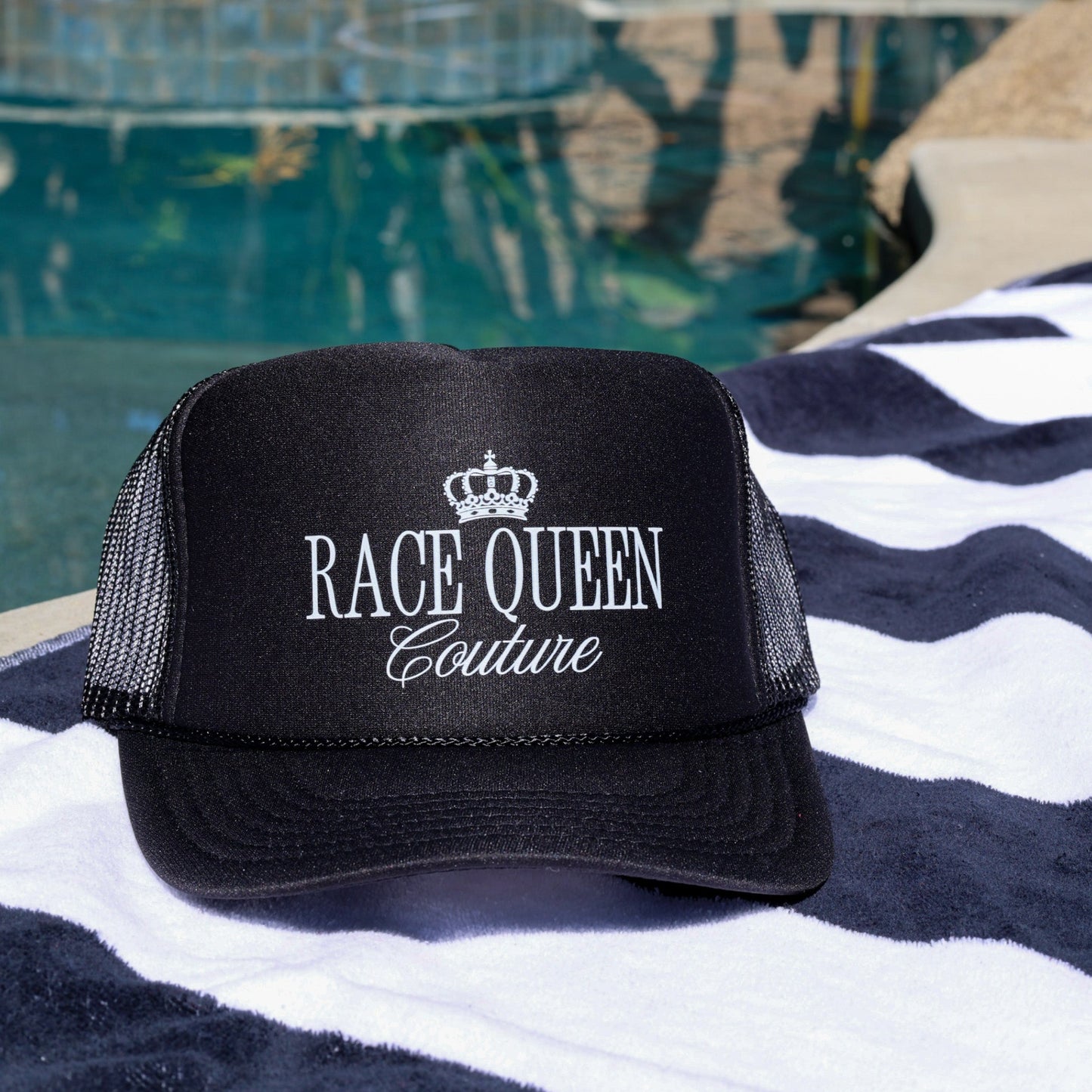 Black cap with 'Race Queen Couture' text on a striped towel by a pool.
